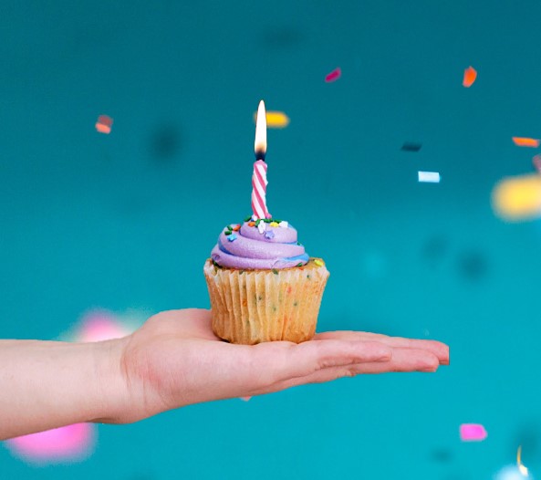 Outstretched hand holding a muffin with one candle in front of a confetti background